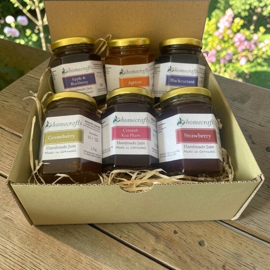 A green cardboard gift box filled with six jars of Homecrafts jam, arranged on a wooden table with a blurred garden background. The jars include Apple & Blackberry, Apricot, Blackcurrant, Gooseberry, Cornish Kea Plum, and Strawberry.