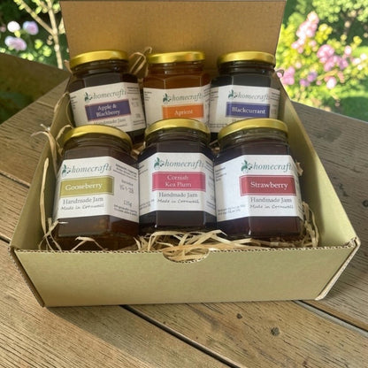 A green cardboard gift box filled with six jars of Homecrafts jam, arranged on a wooden table with a blurred garden background. The jars include Apple & Blackberry, Apricot, Blackcurrant, Gooseberry, Cornish Kea Plum, and Strawberry.