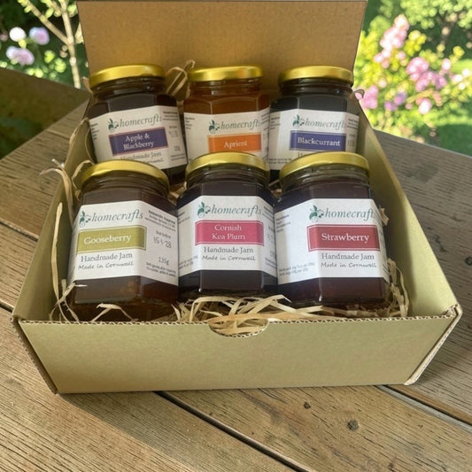 A green cardboard gift box filled with six jars of Homecrafts jam, arranged on a wooden table with a blurred garden background. The jars include Apple & Blackberry, Apricot, Blackcurrant, Gooseberry, Cornish Kea Plum, and Strawberry.